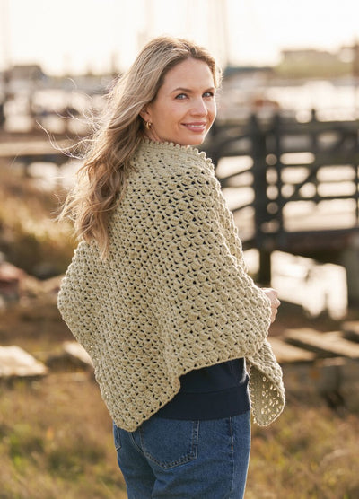 A woman with long hair smiles, wearing a knit shawl by a waterfront. The background features blurred docks and calm water, creating a peaceful tone.