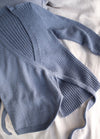 Blue knit wrap sweater with long sleeves and ribbed detail, laid casually on a soft beige fabric. The tone is cozy and relaxed.