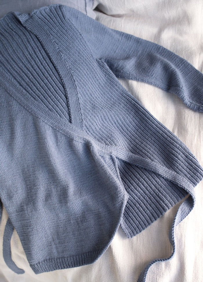 Blue knit wrap sweater with long sleeves and ribbed detail, laid casually on a soft beige fabric. The tone is cozy and relaxed.