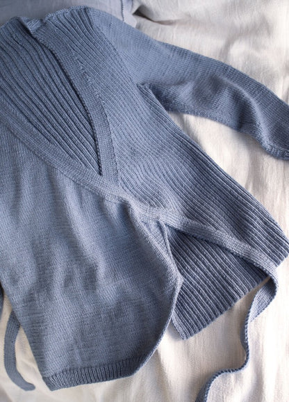 Blue knit wrap sweater with long sleeves and ribbed detail, laid casually on a soft beige fabric. The tone is cozy and relaxed.