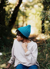 Blue knit cap worn outdoors with a light long-sleeve shirt, surrounded by greenery.