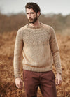 Person wearing the Shire Sweater, a beige knit with intricate chest and sleeve patterns, photographed outdoors among dry foliage.
