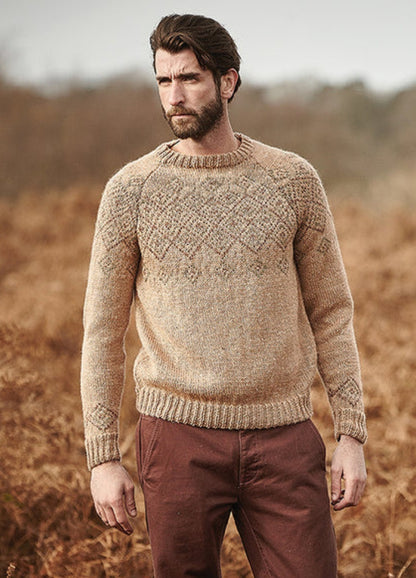 Person wearing the Shire Sweater, a beige knit with intricate chest and sleeve patterns, photographed outdoors among dry foliage.