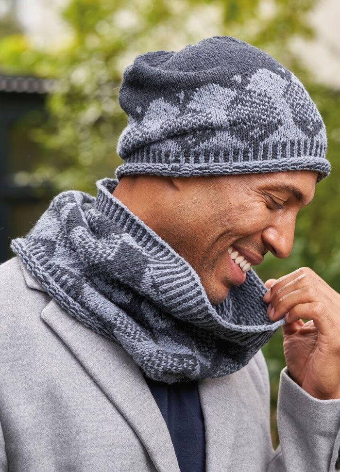 A man wearing a cozy, patterned knit hat and a matching scarf, both in shades of blue and gray, standing outdoors.