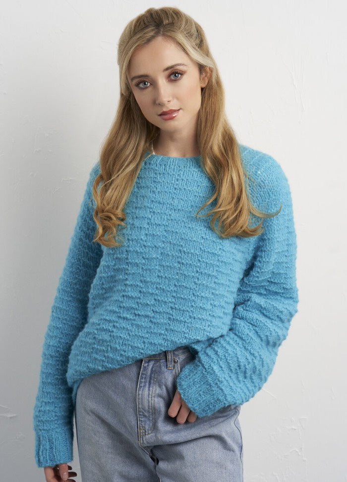 Young woman with long blonde hair wearing a textured blue sweater and light jeans against a white background. She has a relaxed and approachable expression.
