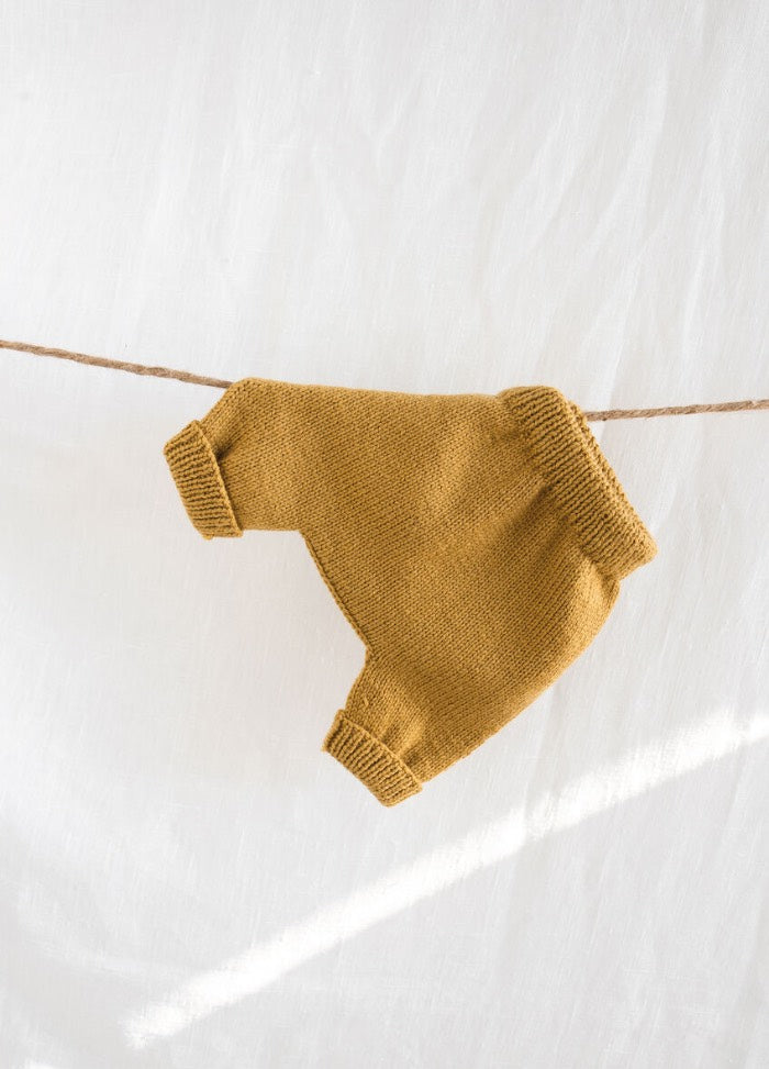 A pair of mustard yellow, knitted children's pants hangs on a string against a white background. The soft texture creates a warm, cozy feel.