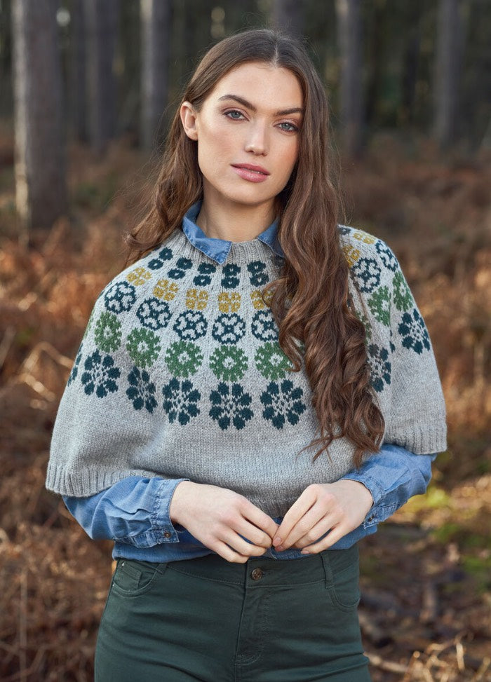Young woman in a forest, wearing a patterned gray sweater over a blue shirt. Her expression is serene, with autumn foliage in the background.