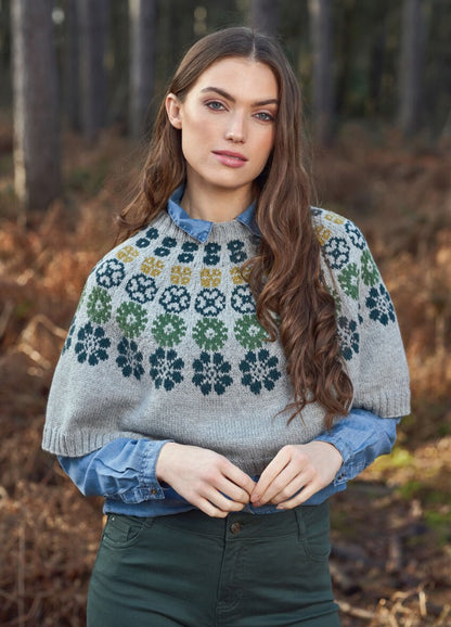 Young woman in a forest, wearing a patterned gray sweater over a blue shirt. Her expression is serene, with autumn foliage in the background.