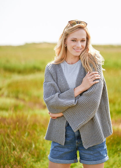 Smiling woman in a gray cardigan and denim shorts stands in a sunlit field. Sunglasses are perched on her head, exuding a relaxed, cheerful vibe.