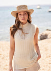 Woman in a straw hat wearing a sleeveless cream knit top stands on a sunny beach with a serene expression. The sea and boats are in the background.