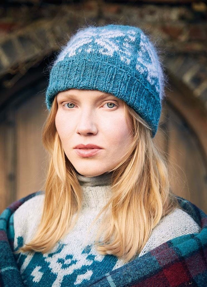A woman wearing a blue knit hat with a white pattern, matching sweater, and a plaid shawl. Her expression is calm, set against a rustic wooden arch.