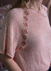 A woman wearing a light pink, knitted top with intricate beadwork in vertical stripes along the shoulder and sleeve, giving a delicate, artistic feel.