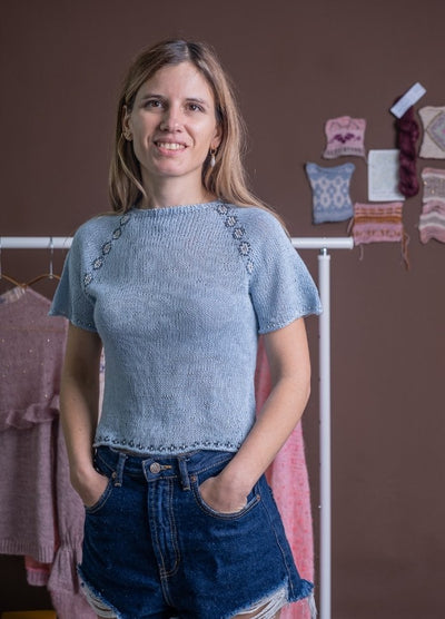Woman in a light blue knit top and denim shorts stands confidently against a brown background. Behind her, knit samples and garments are displayed.