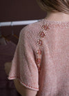 A close-up of a person wearing a pink knitted top featuring an intricate bead design along the shoulder. Soft lighting creates a cozy, delicate tone.
