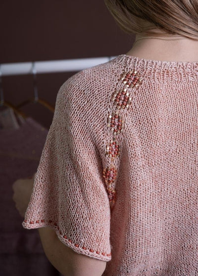 A close-up of a person wearing a pink knitted top featuring an intricate bead design along the shoulder. Soft lighting creates a cozy, delicate tone.