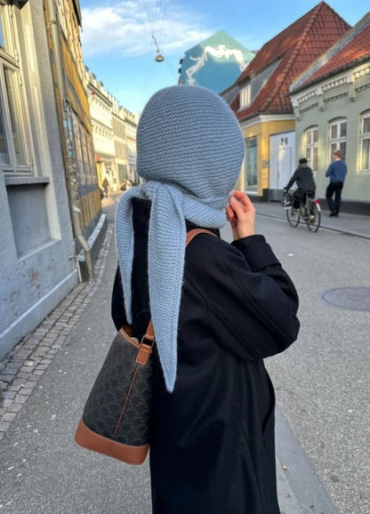 Person in a black coat and blue Sophie Hood stands on a city street. Bicyclist and passerby in the background.