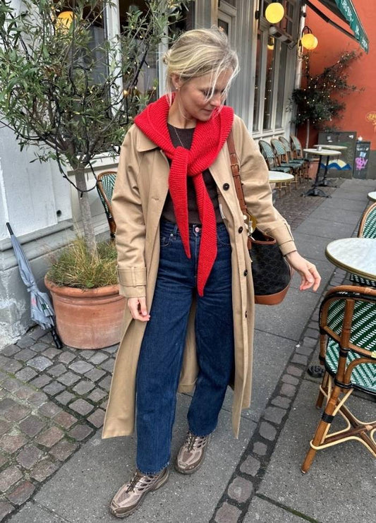 Woman in a beige trench coat with a red Sophie Hood looks down, standing on a cobblestone street with outdoor café tables. Casual, autumn feel.