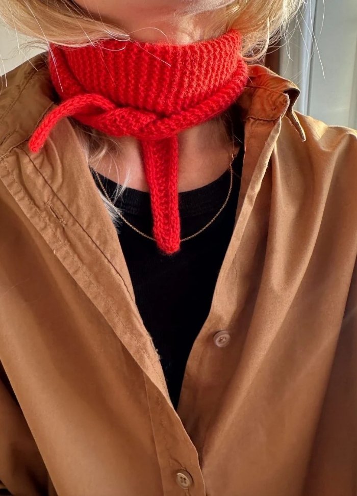 A person wearing a red knit neck scarf tied in a bow over a brown button-up shirt. A glimpse of a black top and a delicate gold necklace is visible.