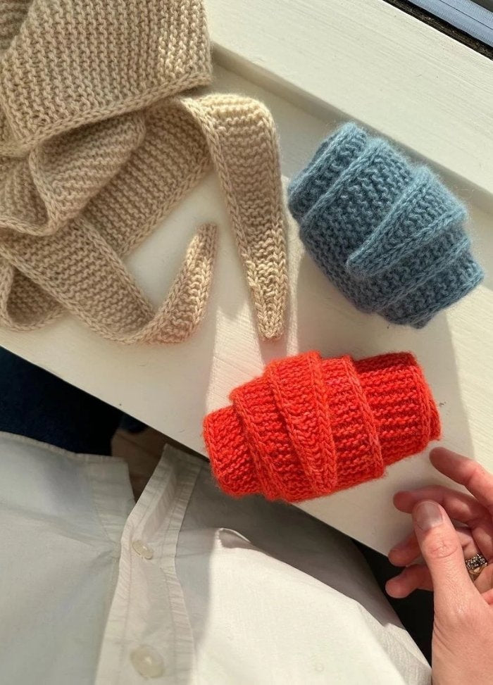 Three knitted scarves in beige, blue, and red are rolled on a table. A person partially visible in a white shirt gently touches the red scarf.