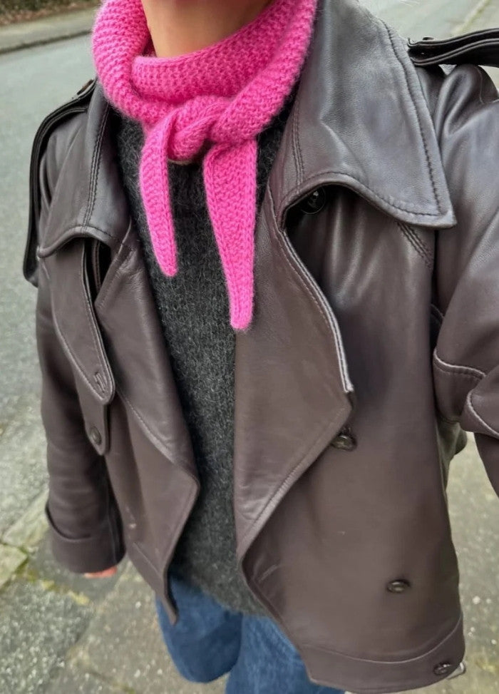 Person wearing a brown leather jacket, grey sweater, and a bright pink scarf standing on a sidewalk. The mood is casual and stylish.