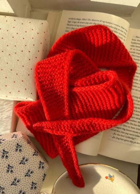 A vibrant red knitted scarf is elegantly draped over an open book with text. Surrounding it are fabric-covered items in white and blue patterns, creating a cozy, warm atmosphere.