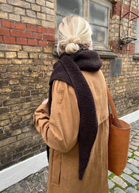 A person with blonde hair in a bun wears a tan coat and brown scarf, carrying a brown tote bag. They face a brick wall and cobblestone path, creating a cozy, autumnal feel.