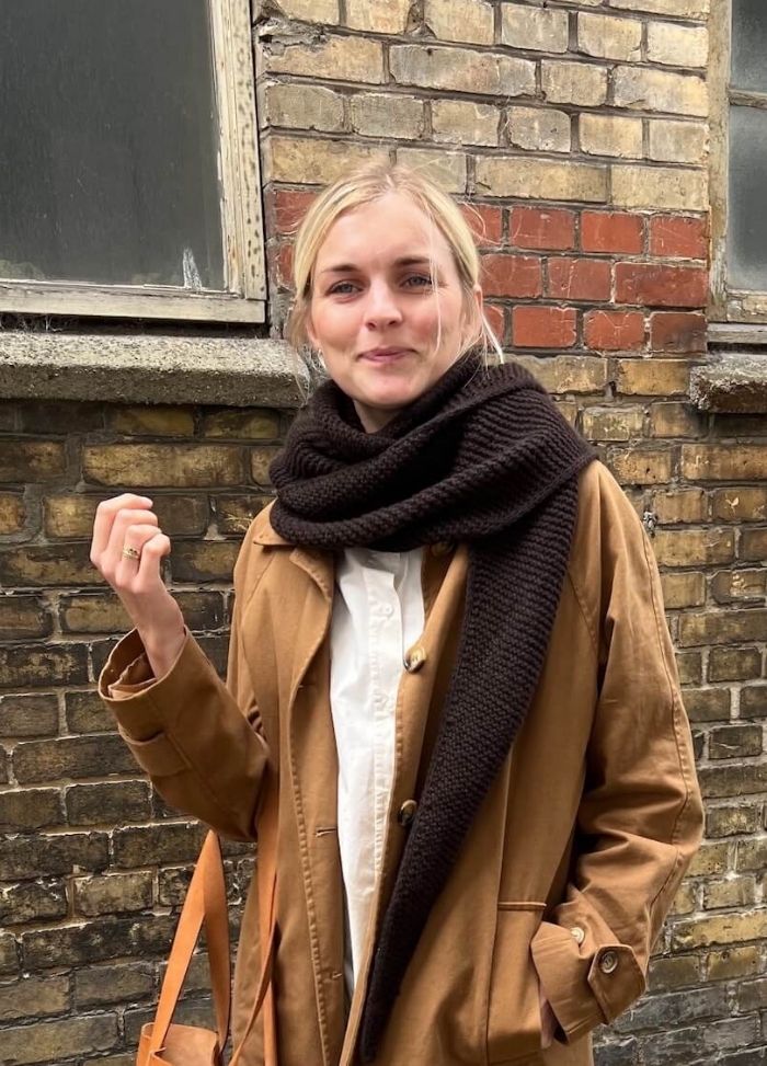A person in a tan coat and dark scarf stands in front of a brick wall, smiling slightly. Overcast lighting and casual atmosphere convey a relaxed mood.