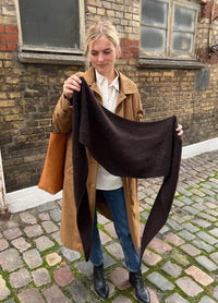Woman standing on a cobblestone street, holding a dark knitted scarf. She wears a tan coat, jeans, and black boots, with a calm expression. Brick wall in the background.