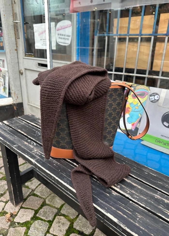A brown knit scarf drapes over a designer handbag on a worn black bench. The scene is outside, with cobblestone pavement and storefronts in the background.