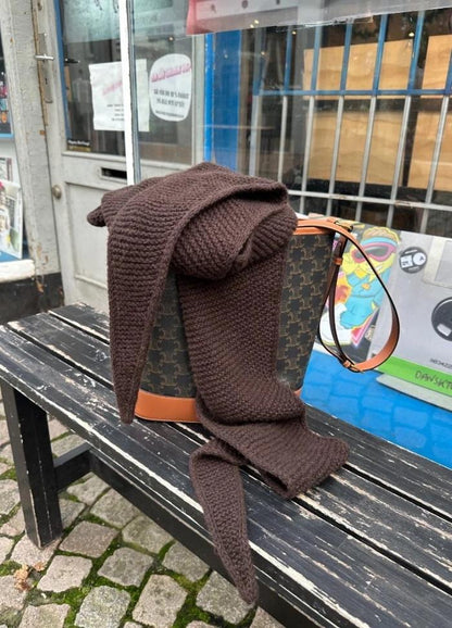 A brown knit scarf drapes over a designer handbag on a worn black bench. The scene is outside, with cobblestone pavement and storefronts in the background.