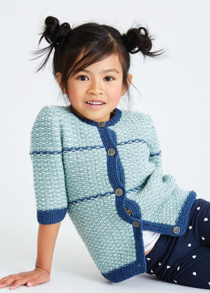 Young girl with two buns in her hair smiles, wearing a blue knitted cardigan with buttons and navy pants with white hearts. She is sitting casually.