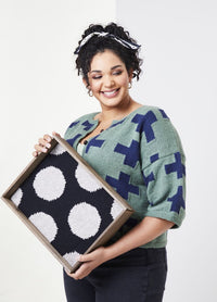 Smiling woman with curly hair holds a wooden frame displaying a black fabric with large white circles. She wears a green sweater with blue crosses.