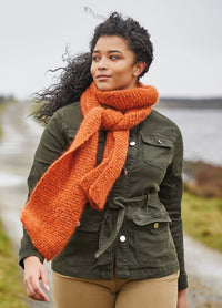 A person walks casually along a coastal path, wearing a green jacket and a large orange scarf. Their curly hair is blowing in the wind, creating a relaxed, contemplative mood.