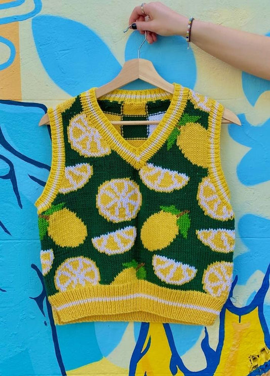A vibrant knit vest with yellow lemons on a dark green background is displayed against a colorful mural. A hand holds the hanger, creating a lively mood.