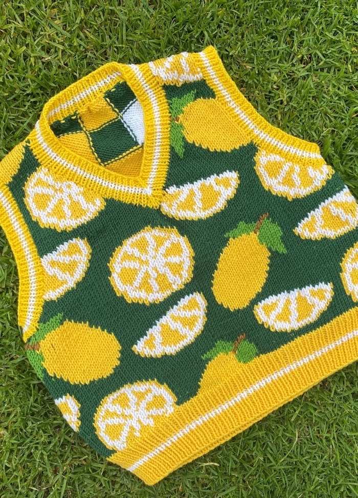 Knitted green vest with yellow and white lemon patterns. It features whole lemons and slices, set against grass, conveying a cheerful, sunny vibe.