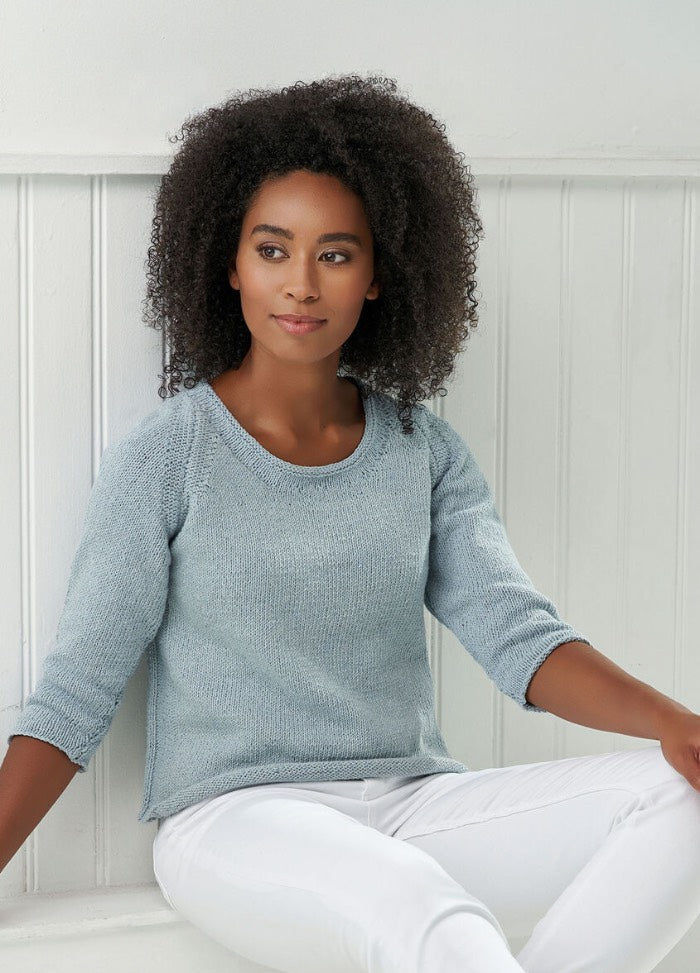 A person with curly hair sits on a white bench, wearing a light blue knitted sweater and white pants. They appear relaxed, looking to the side thoughtfully.