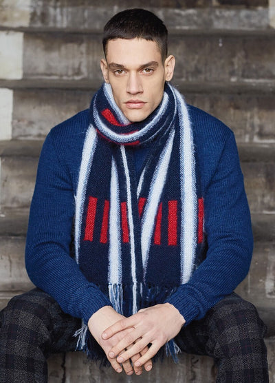 A person in a blue sweater and plaid pants sits on stone steps. They wear a blue, red, and white striped scarf, with a serious expression.