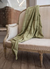 Elegant beige armchair with ornate wood framing on aged wooden floor. A soft, sage green knit blanket is draped casually over it, evoking coziness.