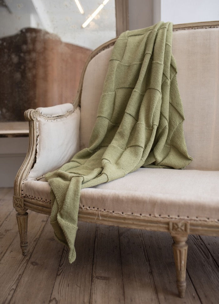 Elegant beige armchair with ornate wood framing on aged wooden floor. A soft, sage green knit blanket is draped casually over it, evoking coziness.
