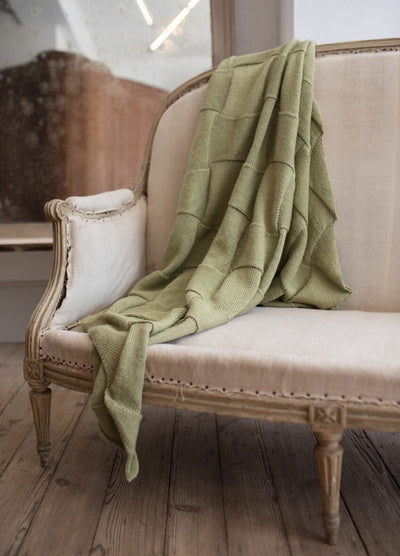 Elegant beige armchair with ornate wood framing on aged wooden floor. A soft, sage green knit blanket is draped casually over it, evoking coziness.