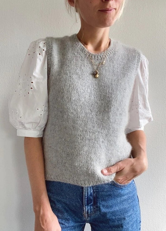 A person wears a grey knit vest over a white puff-sleeve blouse with eyelet details, paired with blue jeans. The casual outfit is accessorised with layered necklaces.