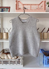A grey knitted sleeveless sweater hangs on a black hanger against a white shelf. The shelf holds beige containers, a pink basket, and a framed photo.