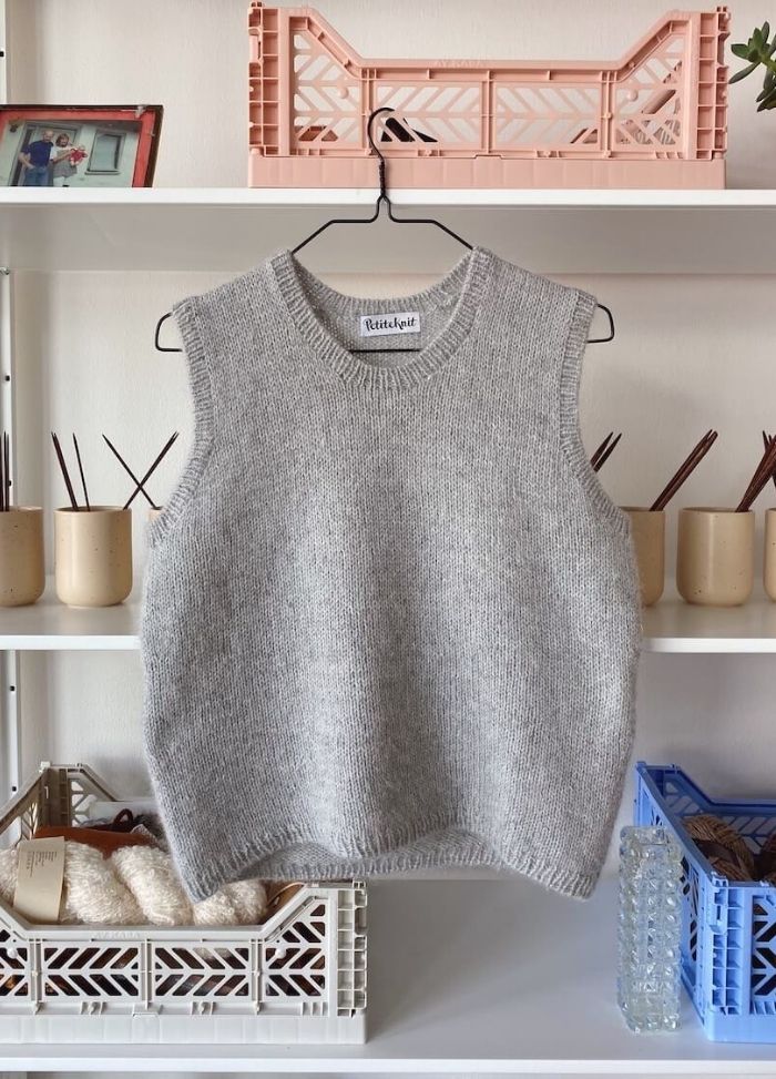 A grey knitted sleeveless sweater hangs on a black hanger against a white shelf. The shelf holds beige containers, a pink basket, and a framed photo.