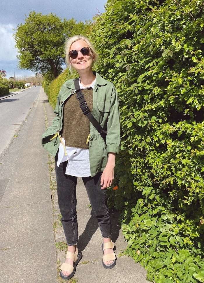 A person stands smiling on a sunny sidewalk, wearing sunglasses, a green jacket, and sandals. Lush greenery lines the path, creating a cheerful vibe.