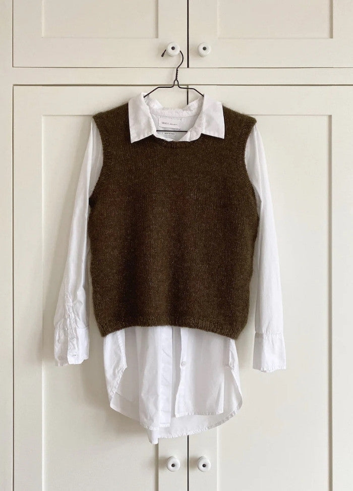 A brown knitted vest layered over a white collared shirt hangs on a hanger against a white panelled wardrobe. The scene conveys a minimalist and cozy style.