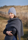 Woman in a knitted blue hat and gray shawl stands outdoors on a sunny day. The background is a rural landscape with a wooden fence. The tone is serene.