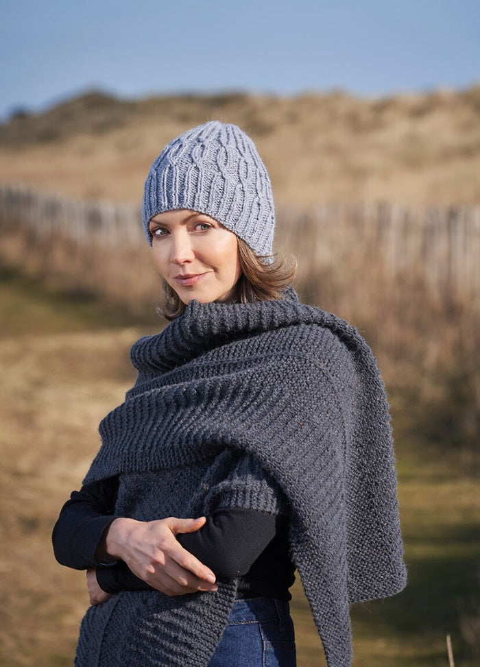 Woman in a knitted blue hat and gray shawl stands outdoors on a sunny day. The background is a rural landscape with a wooden fence. The tone is serene.
