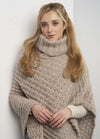 A woman with long blonde hair in a cozy beige knit sweater with a high collar and intricate pattern, leans against a white wall, conveying warmth and comfort.