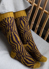 70s Funk Socks: yellow socks with purple wavy abstract patterns, shown seated beside a wicker chair.