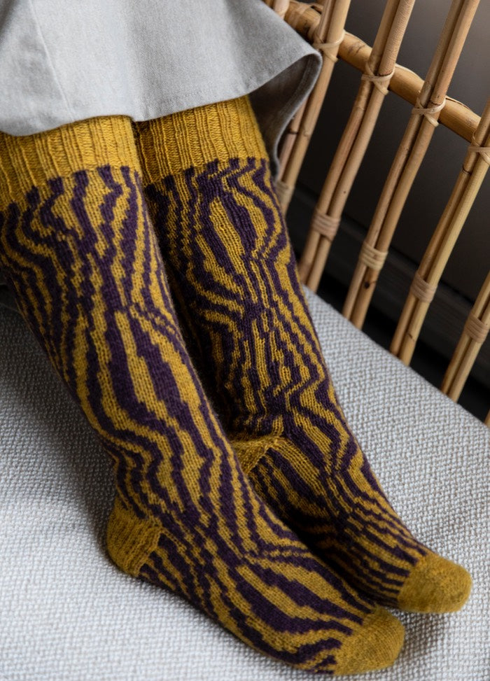 70s Funk Socks: yellow socks with purple wavy abstract patterns, shown seated beside a wicker chair.